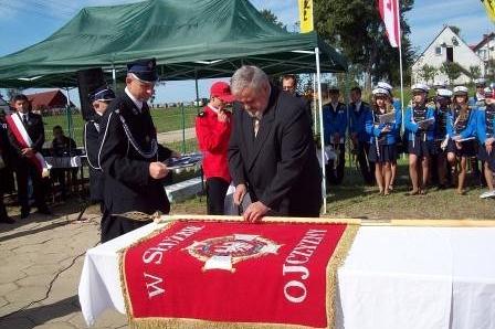 Osoby przy stole z czerwonym sztandarem OSP Lewino na plenerowej uroczystości.