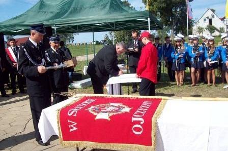 Ceremonia przekazania sztandaru OSP Lewino z czerwonym banerem na stole i uczestnikami.
