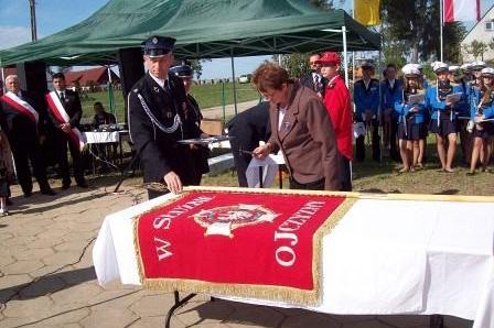 Sztandar OSP Lewino na stole podczas ceremonii, wokół umundurowani goście i zielony namiot.