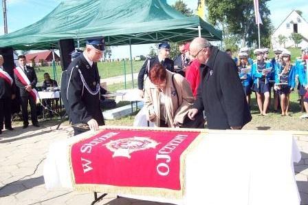 Ludzie podczas ceremonii przekazania sztandaru OSP Lewino, na stole fragment czerwonego sztandaru.