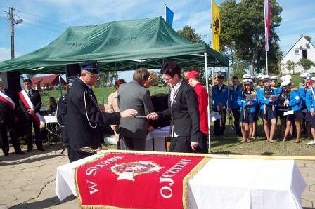 Sztandar OSP Lewino na stole podczas uroczystości z oficjelami i strażakami pod namiotem.