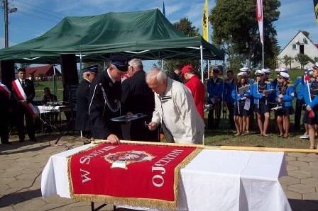 Uroczystość przekazania sztandaru OSP Lewino z ludźmi w mundurach i sztandarem na stole.