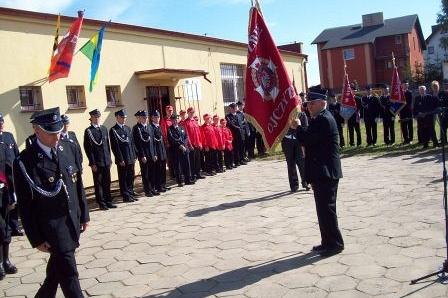 Uroczystość OSP Lewino: strażak w mundurze prezentuje sztandar przed zgromadzoną jednostką.