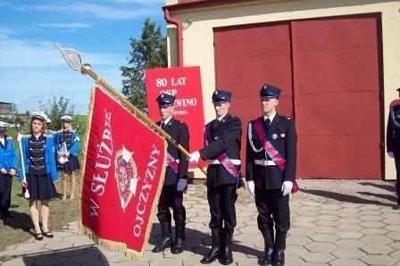Młodzież w galowych mundurach z czerwonym sztandarem OSP Lewino przed remizą strażacką.