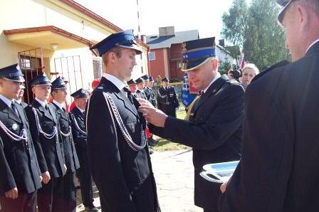 Starszy druh przypina odznakę młodszemu strażakowi podczas ceremonii OSP Lewino.