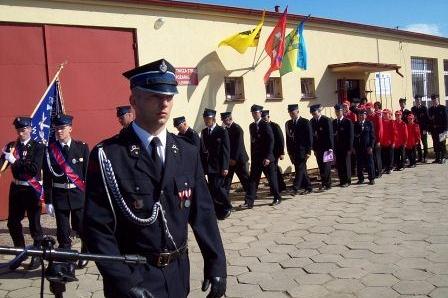 Strażak OSP Lewino w galowym mundurze prowadzi defiladę ze sztandarem przed budynkiem remizy.