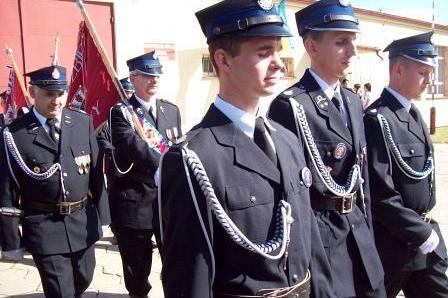 Młodzież OSP w galowych mundurach z białymi sznurami, w tle maszeruje z czerwonym sztandarem.