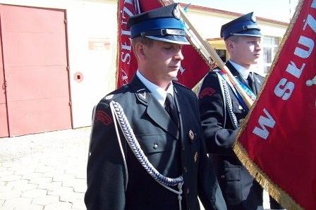Strażacy OSP w galowych mundurach niosą czerwone sztandary podczas uroczystości.