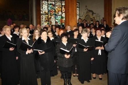 Chór w czerni śpiewa requiem, dyrygent stoi tyłem do widza w sali z witrażami.
