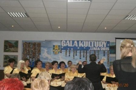 Chór śpiewa pod dyrekcją prowadzącego na tle baneru Gali Kultury.