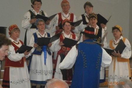 Chór w regionalnych strojach śpiewa z nut, prowadzony przez dyrygenta.