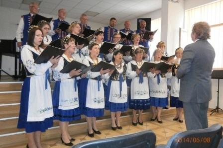 Chór ubrany w stroje ludowe występuje na scenie w Domu Kultury pod batutą dyrygenta.
