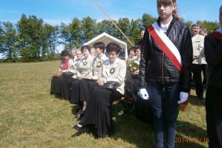 Kobieta z szarfą przed chórem śpiewającym pod namiotem na trawie podczas występu w Lubocinie.