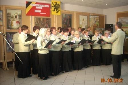 Występ chóru w GDK Linia: dyrygent prowadzi śpiewaków w jasnych strojach z nutami.