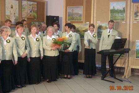 Chór GDK Linia z muzykiem przy keyboardzie, trzymający kwiaty, w tle obrazy.