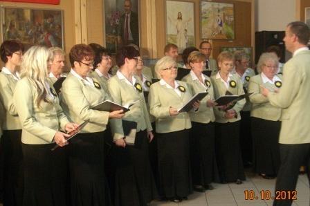 Chór kobiet w jasnych strojach z dyrygentem podczas koncertu w GDK Linia.