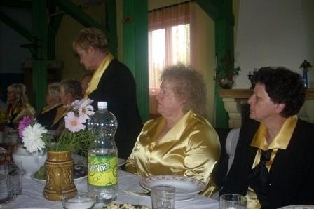 Uczestniczki dożynek przy stole, jedna stoi, dwie siedzą, w ciemnych marynarkach z żółtym.