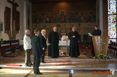 Grupa osób, w tym chórzyści lub duchowni, w kościele przed ołtarzem i amboną.