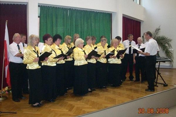 Chór Pięciolinia występuje na scenie, śpiewając z nut pod batutą dyrygenta.