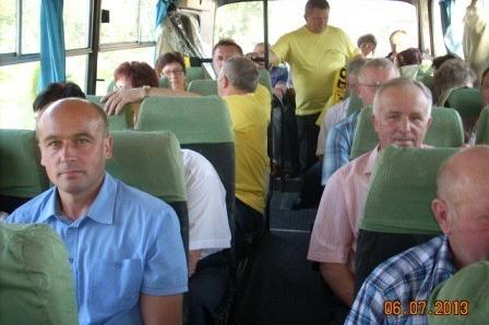 Pasażerowie w autobusie na zielonych siedzeniach, jeden stoi, wielu patrzy w stronę kamery.