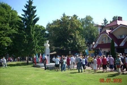 Uczestnicy Zjazdu Kaszubów gromadzą się na trawniku przy pomniku, w tle drzewa.