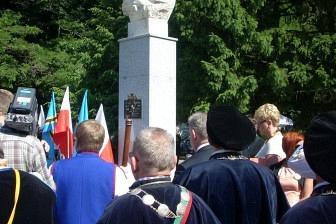 Oficjalna delegacja Kaszubów w strojach galowych przed pomnikiem wśród zieleni.