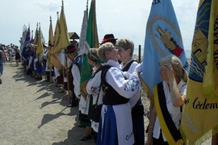 Procesja Kaszubów w tradycyjnych strojach z barwnymi flagami.