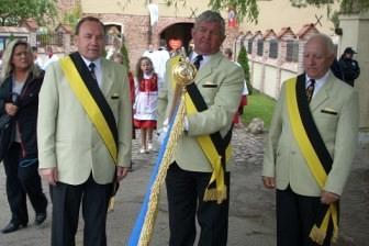 Delegacja mężczyzn w galowych mundurach z szarfami i proporcem na tle historycznych budynków Żukowa.