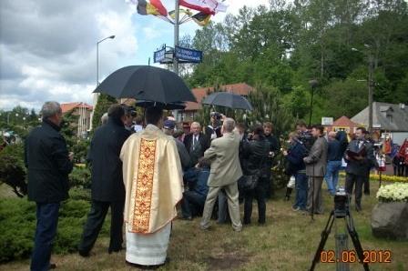 Plenerowa uroczystość w Żukowie z udziałem duchownego i wielu osób pod parasolami.