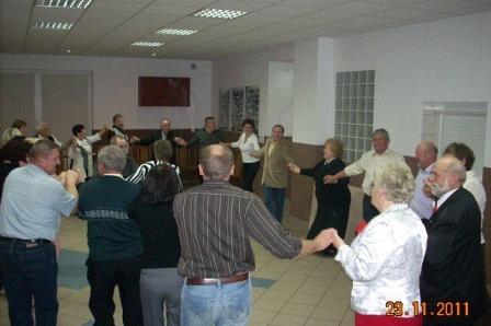 Grupa osób tańczy w kręgu, świętując Andrzejki w przestronnej sali.