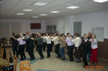 Grupa osób tańczy razem w długiej linii podczas zabawy andrzejkowej w sali.