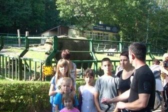 Grupa osób, w tym dzieci, na wycieczce krajoznawczej w Gdańsku, w zielonym otoczeniu.