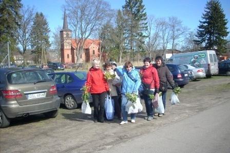 Uczestnicy Targów Ogrodniczych z zakupami na tle parkingu i drzew w Srzelinie.