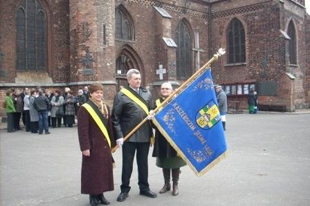 Trzy osoby w ceremonialnych strojach prezentują kaszubską flagę na tle zabytkowego budynku.