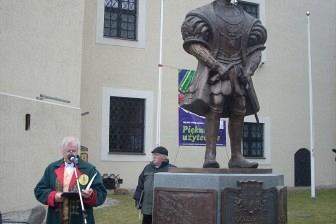 Statua Kaszuba w stroju ludowym obok małej postaci trzymającej różowy przedmiot.