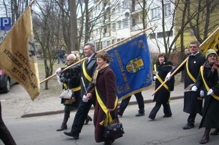 Grupa osób w uroczystych strojach maszeruje z kaszubską flagą i sztandarami.