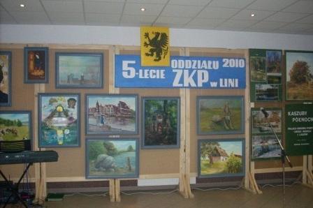 Wystawa obrazów na drewnianych sztalugach z bannerem 5 lat ZKP i emblematem.