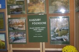 Ekspozycja jubileuszowa ZKP O/LINIA z panelami i zdjęciami na drewnianych stojakach, z kwiatami.