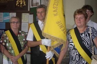 Trzy osoby z żółtymi szarfami i proporcem na ceremonii odsłonięcia pomnika.