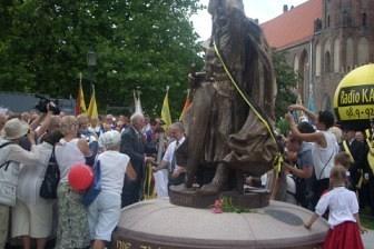 Pomnik Świętopełka w Gdańsku otoczony ludźmi na placu, z budynkiem w tle.