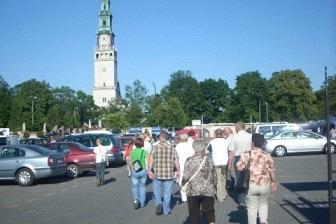 Uczestnicy wycieczki idą parkingiem obok aut, w tle błękitne niebo i wysoka wieża.