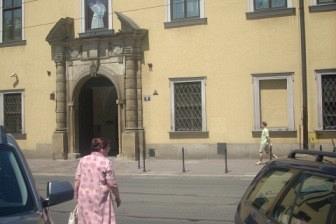 Osoba w różu idzie ulicą obok zabytkowej żółtej kamienicy, między zaparkowanymi samochodami.