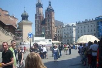 Słoneczny Rynek Główny w Krakowie z Kościołem Mariackim, ludźmi i ulicznymi straganami.