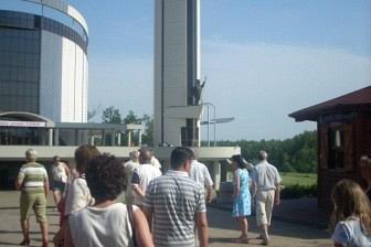 Grupa ludzi idzie pod wysoką wieżą i okrągłym budynkiem w słoneczny dzień.
