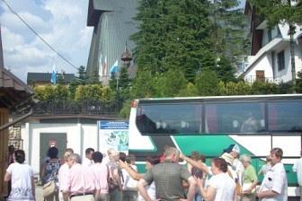 Grupa ludzi pod zabytkowym kościołem, otoczonym drzewami, z widocznym autobusem wycieczkowym.