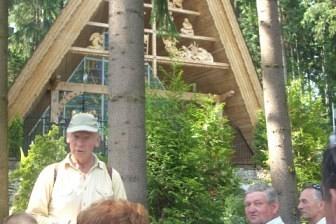 Uczestnicy wycieczki słuchają przewodnika przy drewnianej trójkątnej konstrukcji wśród zieleni.
