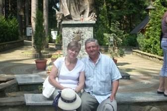 Uśmiechnięta para seniorów na tle rzeźby religijnej i zieleni, podczas wycieczki ZKP.