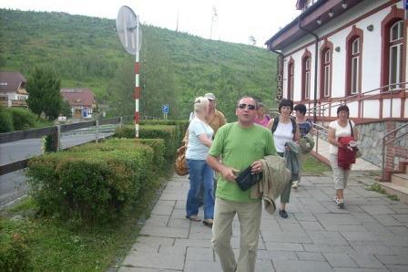Uczestnicy wycieczki idą chodnikiem przy tradycyjnym budynku na tle zielonych wzgórz.