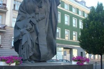 Duża kamienna statua Papieża Polaka na placu miejskim wśród kamienic.