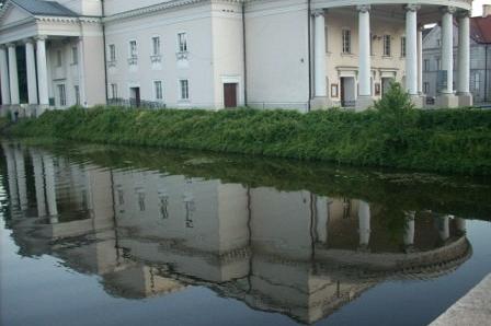 Klasycystyczny budynek z kolumnami i zielenią, odbity w spokojnej tafli wody.